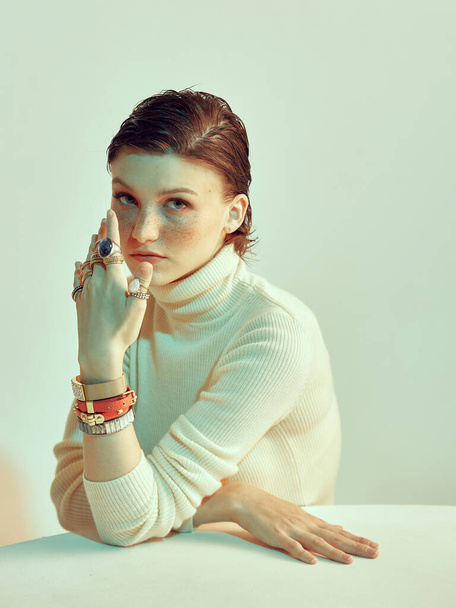 Redhead girl with freckles and stylish short haircut posing in studio. Jewelleries, rings and bracelets on female hand - Zdjęcie, obraz