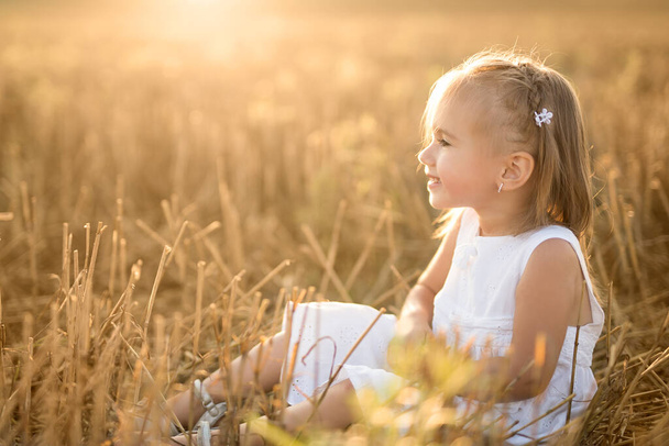  A girl in a white dress sits in a mown wheat field at sunset - Valokuva, kuva