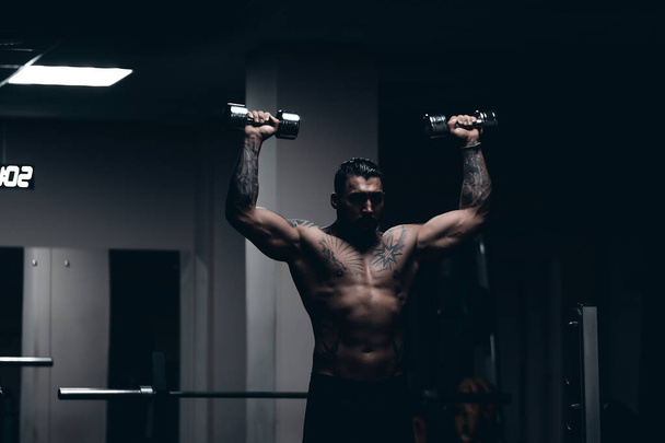 Gespierde mannelijke atleet met getatoeëerde naakte romp staan in donkere sportschool en doen halter krullen tijdens de training - Foto, afbeelding