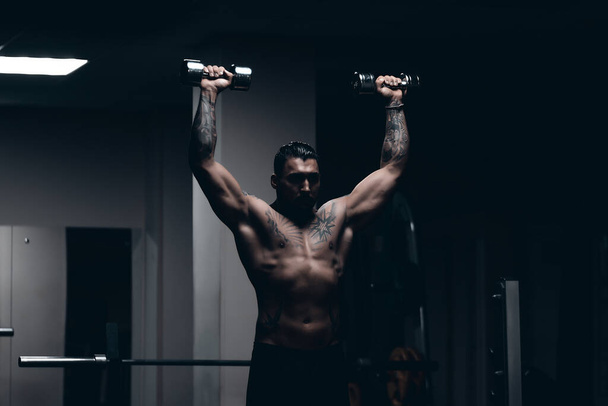 Muskularny sportowiec z wytatuowanym nagim tułowiem stojącym w ciemnej sali gimnastycznej i wykonującym loki hantli podczas treningu - Zdjęcie, obraz