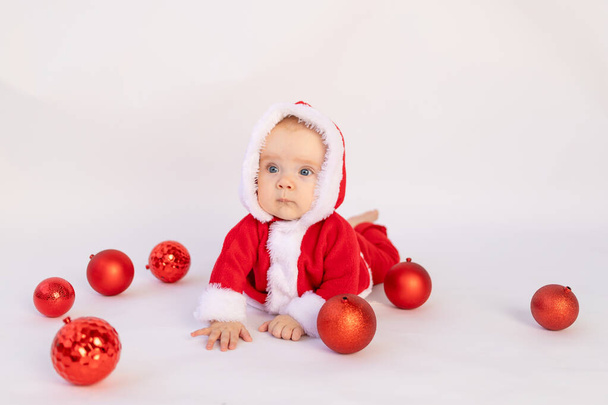 un niño pequeño se encuentra en un traje de Santa sobre un fondo blanco aislado con bolas rojas de Navidad, espacio para el texto, concepto de Navidad - Foto, imagen