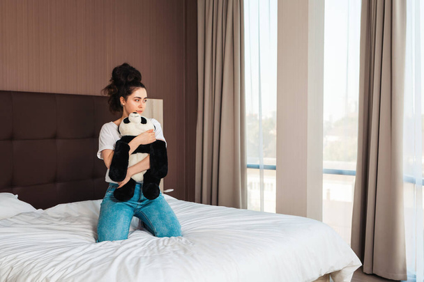 Uma jovem mulher bonita caucasiana abraça um brinquedo de pelúcia em suas mãos enquanto se senta na cama e olha pela janela. Windows em segundo plano. - Foto, Imagem