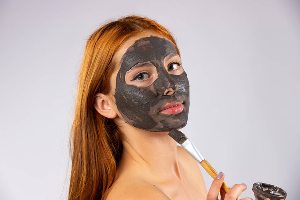  Una joven pelirroja se aplica una máscara de barro en la cara con un cepillo. Foto sobre fondo blanco con espacio lateral vacío. Foto de alta calidad - Foto, Imagen