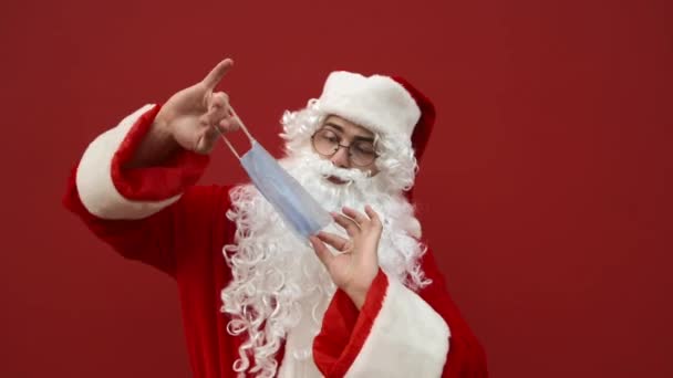 Der lustige Weihnachtsmann hält sich eine Schutzmaske auf und wirft sie ihm angewidert ins Gesicht. Der Weihnachtsmann mag keine Schutzmasken. Der Weihnachtsmann in einer Coronavirus-Pandemie. - Filmmaterial, Video