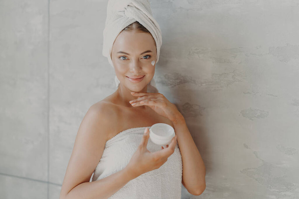 Indoor shot of happy young European woman applies anti aging cream on face, disfruta del tratamiento facial después de tomar la ducha, usa toalla de baño en la cabeza y alrededor del cuerpo desnudo, posa contra la pared gris - Foto, Imagen