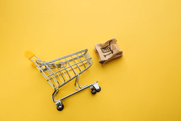 Caja de papel dañada y carrito de compras en una mesa amarilla. Compras en línea, servicio y concepto de entrega - Foto, Imagen