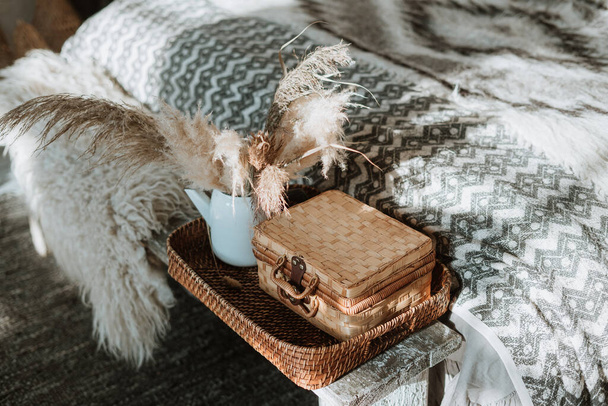 Close-up of wooden bench on carpet against white bed with patterned plaid in bedroom interior with dry glass on table. bohemian chic bedroom - Photo, Image