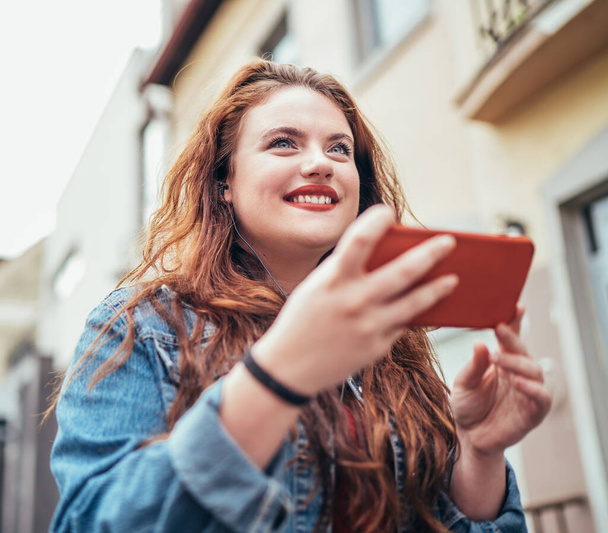 Red curled long hair caucasian teen girl portrait walking on the street and browsing the internet using the modern smartphone. Modern people with technology devices concept image. - Фото, зображення