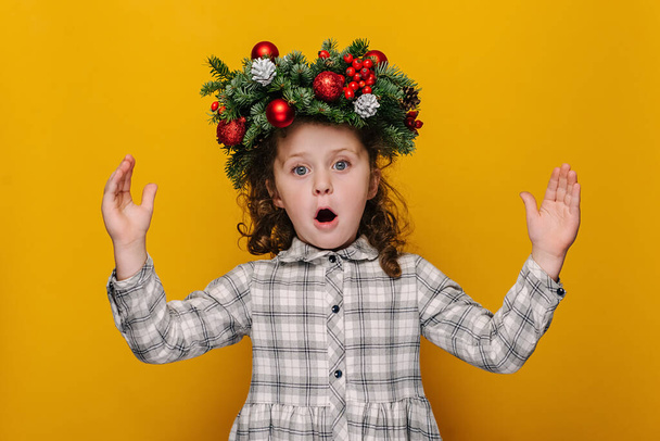 Shocked cute little kid girl showing big size gesture with hands, impressed child demonstrate something wide, wears handmade wreath, isolated on yellow studio background. Merry Christmas concept - Foto, immagini