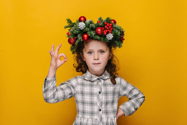 Cute little girl kid in traditional Christmas wreath, makes okay gesture, enjoys life, isolated over pastel yellow studio background, says ok, confirms information. Happy New Year merry holiday - Фото, изображение