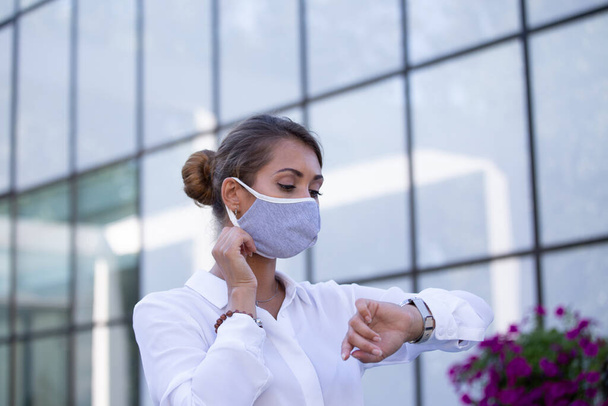 Pretty young business woman with protective mask looking at wristwatch in front of office building. Virus protection and social responsibility concept - Valokuva, kuva
