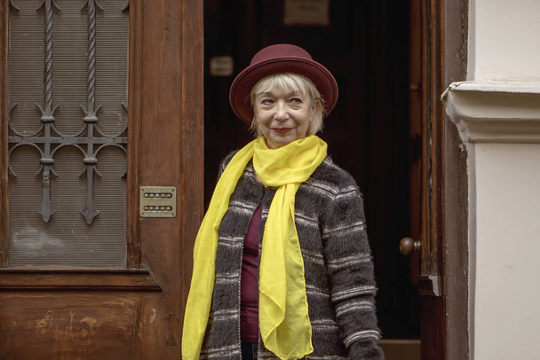 Egy 60-65 éves, kalapos idős nő jön ki egy régi ház bejáratán. Koncepció, nyugdíjazás, divatszakember, kiváló egészség, találkozás az utcán, elszigeteltség és kommunikáció, - Fotó, kép
