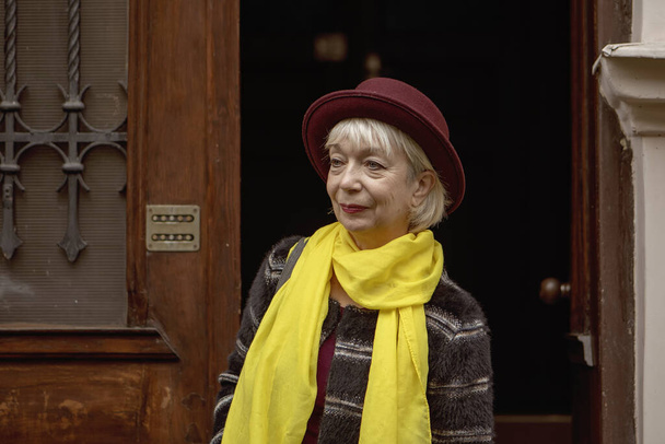 Een oudere vrouw van 60-65 jaar met een hoed komt uit de ingang van een oud huis. Concept, pensioen, fashionista van gevorderde jaren, uitstekende gezondheid, ontmoeting op straat, isolatie en communicatie, - Foto, afbeelding
