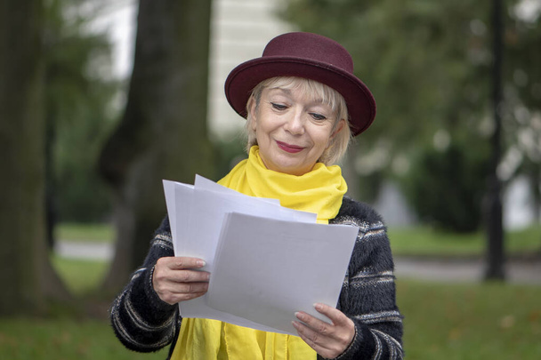Een oudere gepensioneerde 60-65 jaar oud leest documenten met tekst zittend in het park op een bankje. Misschien is ze lerares en controleert ze het werk van studenten of bereidt ze zich voor op een lezing.. - Foto, afbeelding