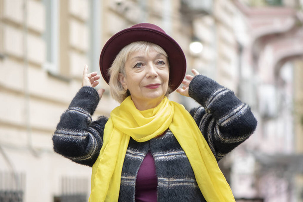 An elderly woman 60-65 years old adjusts a fashionable hat on the background of the city landscape. Concept: retirement, tourism and travel, fashionista of advanced years, excellent health. - Photo, Image