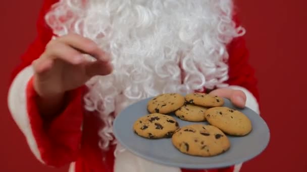 Santa sobre un fondo rojo toma galletas de chocolate de Navidad de un plato. De cerca. - Metraje, vídeo