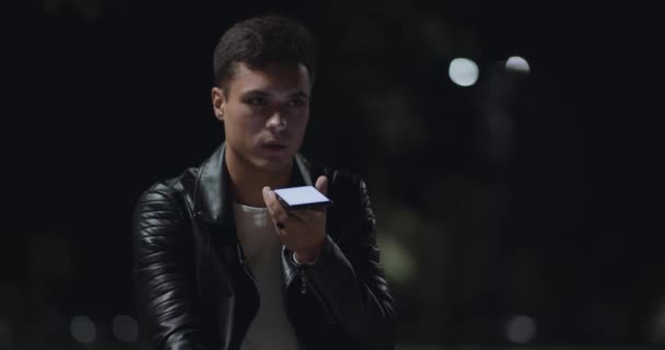 Spracherkennung. Junger ernsthafter Mann nimmt Audio-Botschaft mit Handy auf und sitzt spät abends im Stadtpark - Filmmaterial, Video