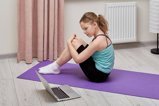 A beautiful cute girl looks at her laptop and plays sports at home on a Mat. - Valokuva, kuva