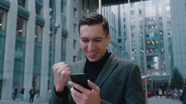 Akıllı telefona bakan mutlu iş adamı çok mutlu hissediyor, mobil başarı iddiasını kutluyor, modern ofis binasının yakınında duruyor. Heyecanlı adam akıllı telefonda online oyun uygulamasını kazanıyor. - Video, Çekim