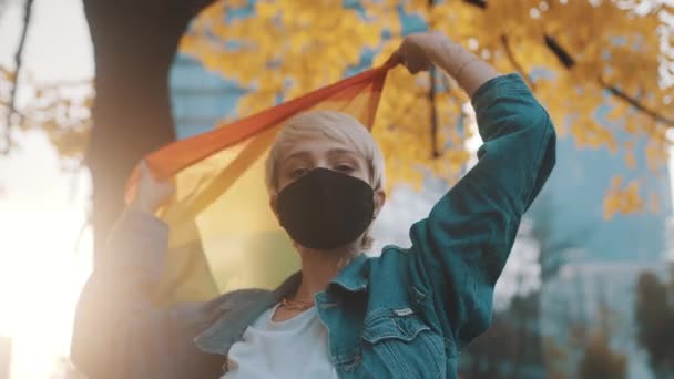 Orgulhoso jovem loira mulher com máscara facial acenando bandeira do arco-íris sob a árvore com folhas amarelas - Filmagem, Vídeo