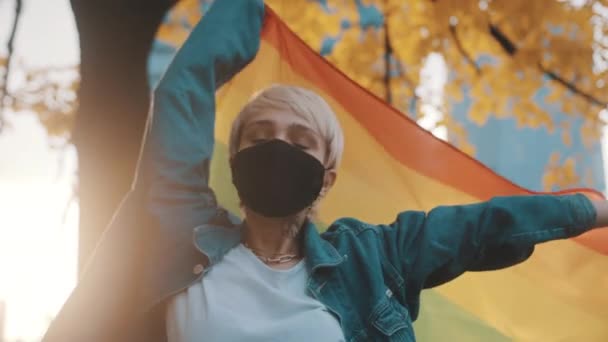 Mulher branca loira nova com máscara facial acenando bandeira do arco-íris sob a árvore com folhas amarelas - Filmagem, Vídeo