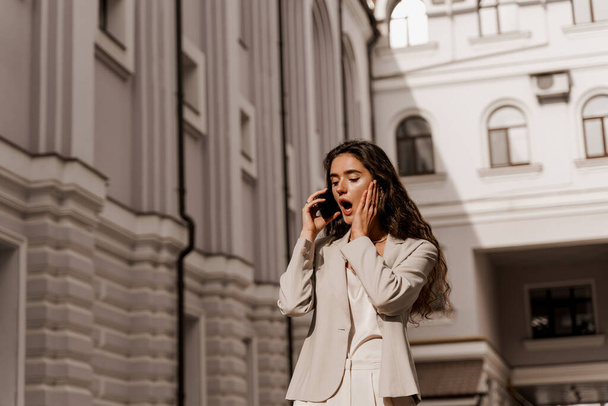 Mujer de negocios hablando por teléfono en la ciudad. Comunicación con amigos. Chica con el pelo rizado vestido con ropa formal blanca - Foto, Imagen