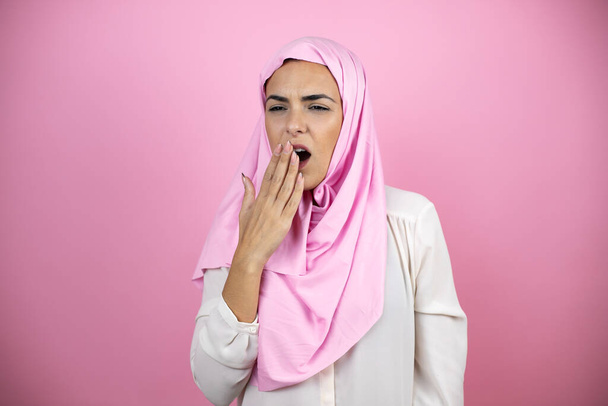 Nuori kaunis arabinainen yllään islamilainen hijab yli eristetty vaaleanpunainen tausta kyllästynyt haukottelu väsynyt peittää suun käsin. Levottomuus ja uneliaisuus. - Valokuva, kuva