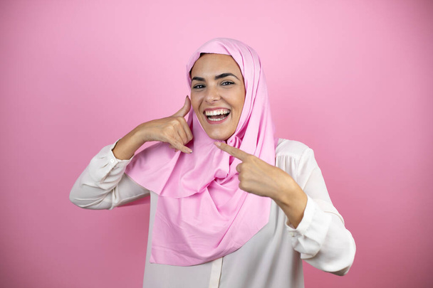 Młoda, piękna arabka, ubrana w islamski hidżab na odizolowanym różowym tle, wykonująca gestykulację rękami. - Zdjęcie, obraz