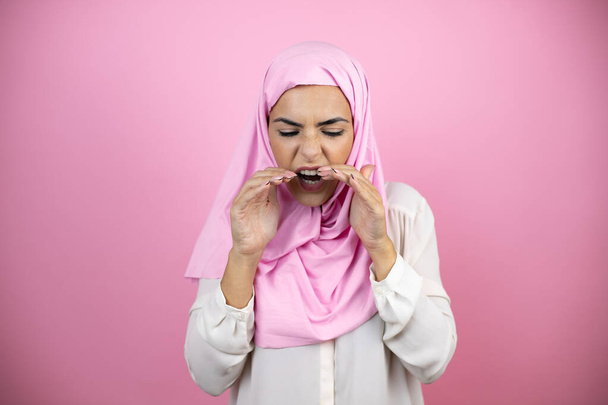 Fiatal gyönyörű arab nő visel iszlám hidzsáb felett elszigetelt rózsaszín háttér kiabál és sikoltozik hangosan le a kezét a száját - Fotó, kép