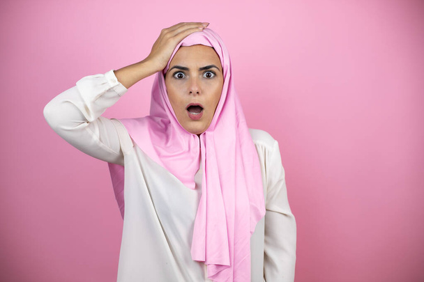 Молода красива арабська жінка в ісламському хіджабі над ізольованим рожевим тлом накладає одну руку на голову посміхаючись, ніби вона щось забула
 - Фото, зображення