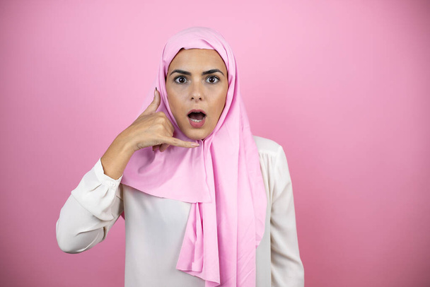 Young beautiful arab woman wearing islamic hijab over isolated pink background confused doing phone gesture with hand and fingers like talking on the telephone - Foto, imagen