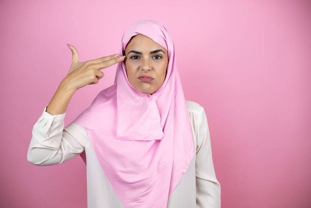 Young beautiful arab woman wearing islamic hijab over isolated pink background Shooting and killing oneself pointing hand and fingers to head like gun, suicide gesture. - Photo, image
