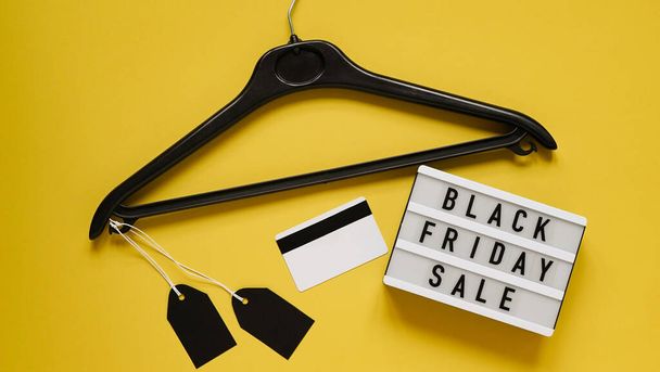 Online shopping for Black Friday sales,pay by credit card. White board with text lettering Black Friday sale,black clothes hanger with black blank tags,credit card yellow background,top view,flat lay. - Valokuva, kuva