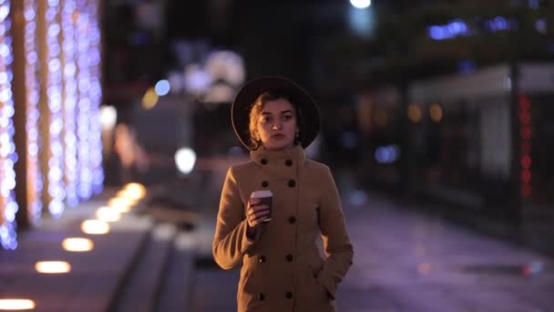Hermosa mujer en sombrero y abrigo bebe café y camina en la ciudad de la noche - Imágenes, Vídeo
