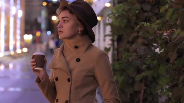 Hermosa mujer en sombrero y abrigo bebe café en la ciudad de la noche - Metraje, vídeo