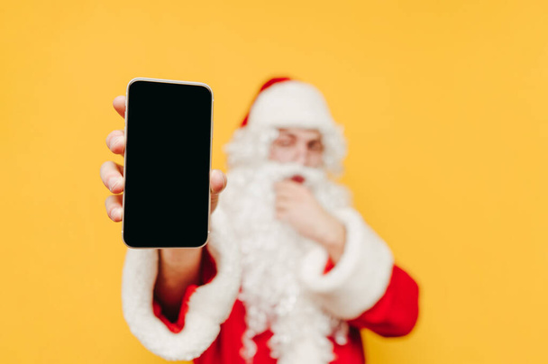 Santa Claus stands on a yellow background, looks at the camera with a pensive face and shows a smartphone with a black screen. Copy space - Photo, image