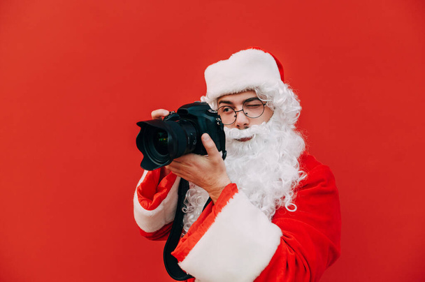 Fotograf Santa Claus stoi na czerwonym tle i robi zdjęcie profesjonalnemu aparatowi z poważną twarzą. Odizolowany. Zdjęcie świąteczne. - Zdjęcie, obraz
