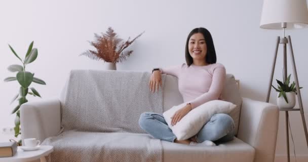 Comfort domestico. Felice donna asiatica seduta sul divano a casa a ridere della macchina fotografica - Filmati, video