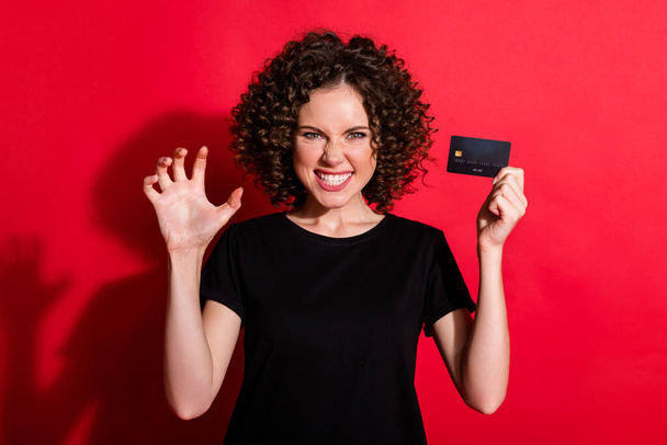Photo d'enfant drôle ondulé dame habillée tenue noire décontractée tenant carte de crédit montrant dents isolées fond de couleur rouge - Photo, image