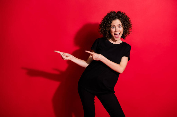 Photo of surprised funny curly woman wear casual black outfit pointing two fingers empty space isolated red color background - Fotoğraf, Görsel