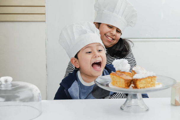 Hermanos latinos con gorras de chef divirtiéndose en la cocina - niños hispanos sonriendo mientras terminan de decorar el pastel - niños cocinando - Foto, imagen