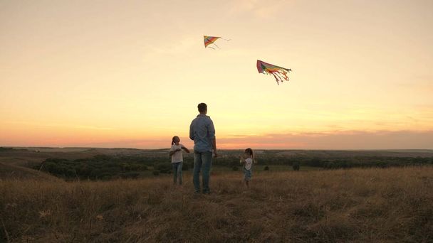 Apa és szeretett lányai színes papírrepülőket indítanak az égbe. Apa gyerekekkel játszik sárkányokkal naplementekor a parkban. egészséges család a szabadban. szórakoztató játszani a szülőkkel a napon - Fotó, kép