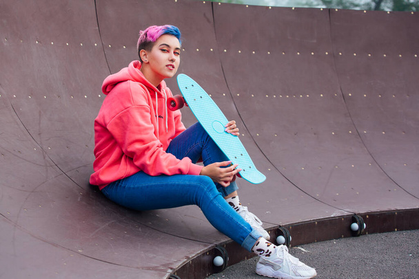 Stylish young woman with short colored hair sitting with her blue plastic skateboard in skatepark. Youth concept. - Foto, Bild