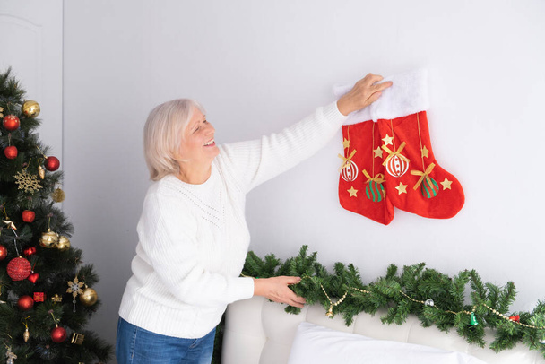 Positieve senior vrouw versiert de kamer voor Kerstmis. levensstijl binnen, ouderdom en vakantie concept. - Foto, afbeelding