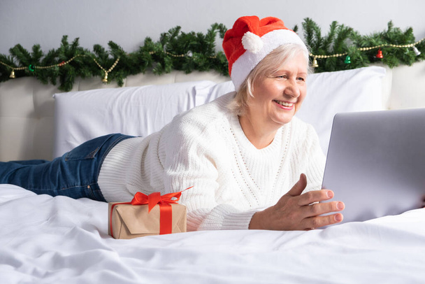 Salutations en ligne de Noël. heureuses femmes âgées dans le chapeau de Père Noël à la maison au lit. style de vie intérieur, technologie, vieillesse et vacances concept. - Photo, image