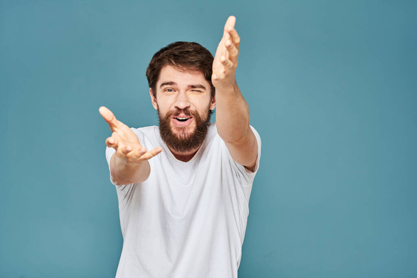Un hombre barbudo con una camiseta blanca gestos con las manos emociones fondo azul - Foto, imagen