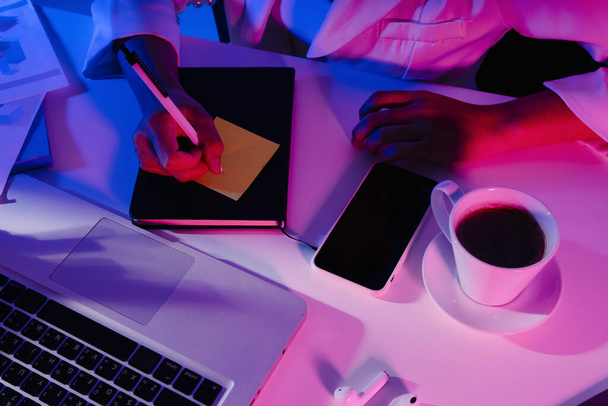 Pohled shora - obchodnice pracovní stůl s notebookem, kniha, káva, telefon v neonové barevné světlo. - Fotografie, Obrázek