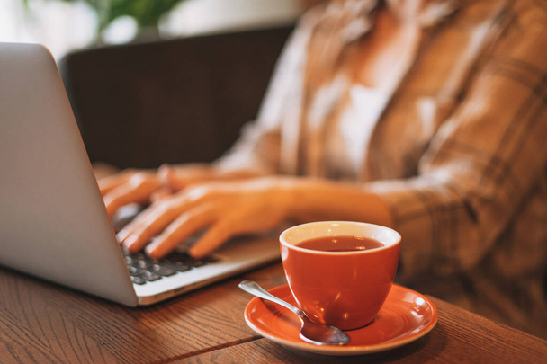 Rode kop thee op de achtergrond van de vrouw in geruite shirt werken met laptop in cafe - Foto, afbeelding