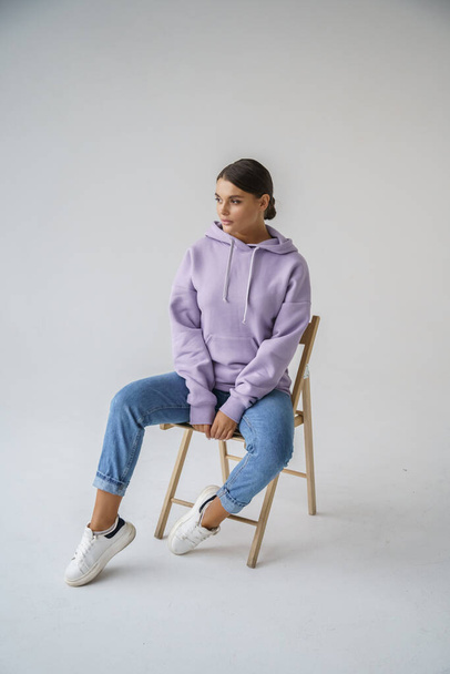 Portrait of beautiful young brunette in purple hoody and blue jeans posing on gray studio background - Foto, Imagen