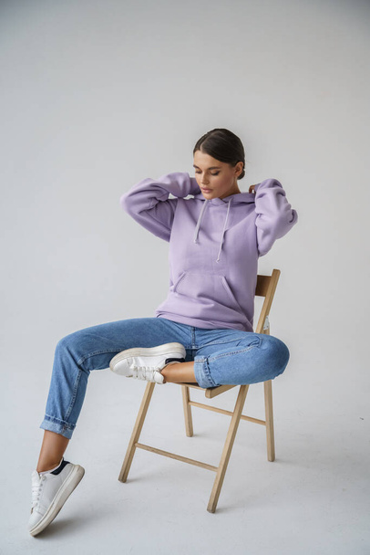 Portrait of beautiful young brunette in purple hoody and blue jeans posing on gray studio background - Photo, Image